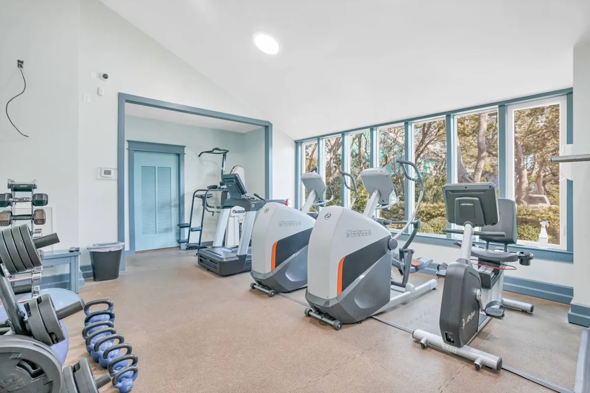 A bright, well-equipped fitness room featuring elliptical machines, stationary bikes, and dumbbells. The space is clean and spacious, ideal for workouts and personal fitness routines while on vacation.