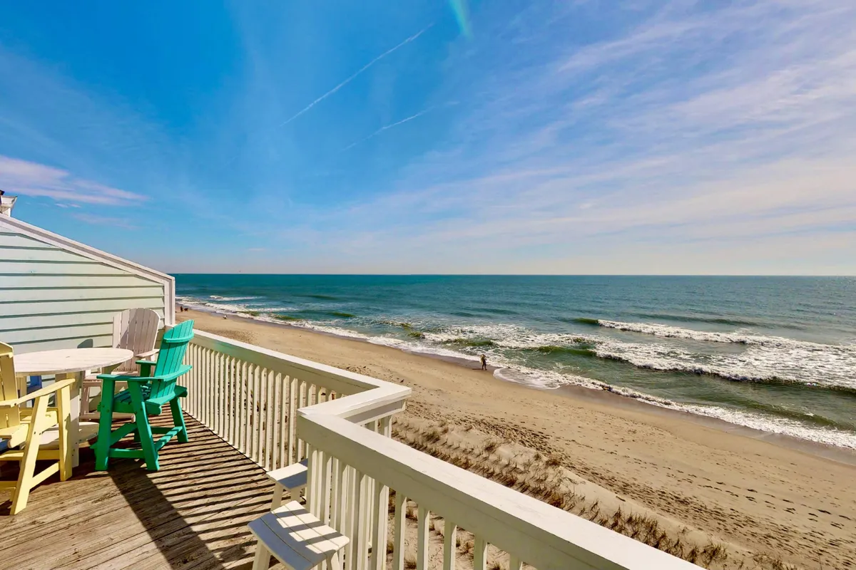 The image showcases a sun-kissed oceanfront deck furnished with inviting outdoor seating, overlooking sweeping views of the sparkling Atlantic and a pristine stretch of sandy beach. The tranquil scene suggests activities like dining outdoors, lounging in the sun, or strolling along the shoreline, all set beneath a wide, serene sky.