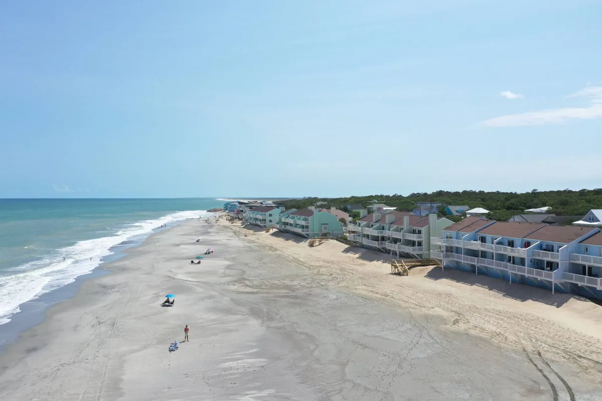 A lively beachfront scene with families and groups enjoying the sandy shore, calm ocean waves, and wide stretches of sandy beach. Children are building sandcastles, people are walking along the water, and the overall mood is carefree and inviting. Beachfront properties frame the background, showcasing direct access to the shore.