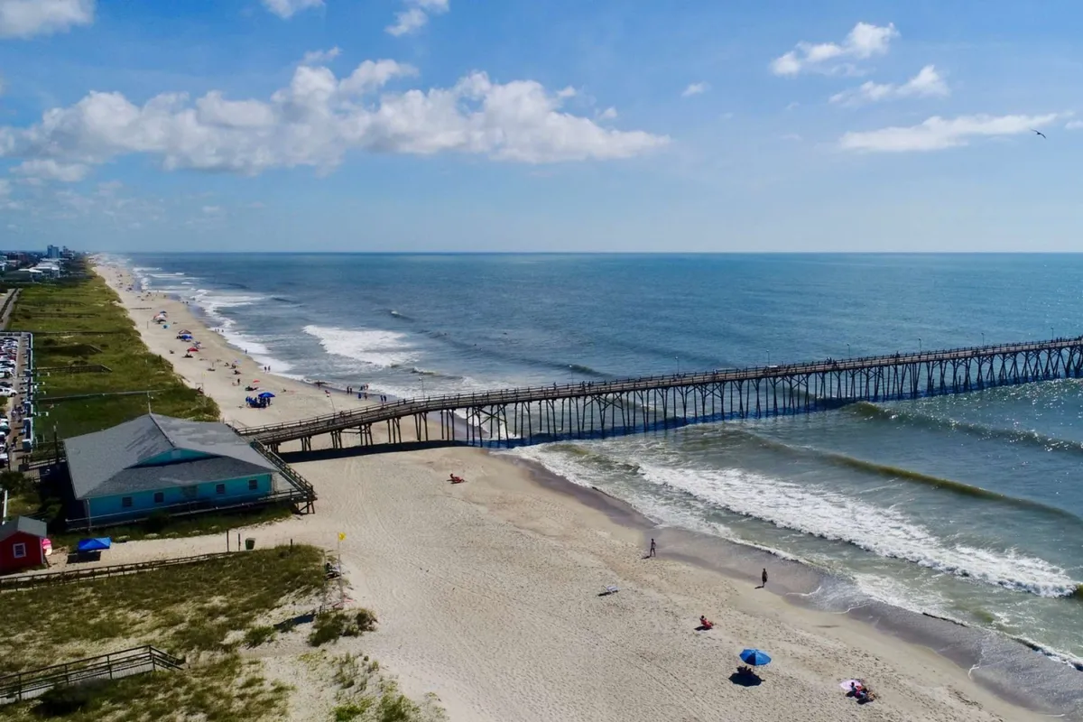 A lively pier extends over the ocean, with people enjoying activities like fishing, swimming, and strolling along the beach. Colorful umbrellas and sunbathers dot the sandy shore, creating an inviting and carefree mood. The pier itself is a clear nod to local fishing hotspots mentioned in the content.