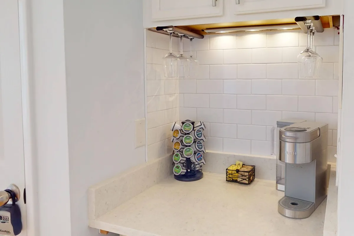 A cozy coffee bar setup with a Keurig machine, cup holder, hanging glasses, and white subway tile backsplash. The scene is designed for both functionality and a touch of style, capturing the essence of morning routines and social moments centered around coffee.