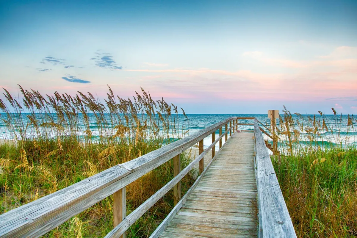 A tranquil wooden boardwalk leads through beach grass toward the ocean, with gentle waves and a colorful sunset sky in the background. The scene exudes peace and natural beauty, inviting leisurely strolls and quiet reflection.