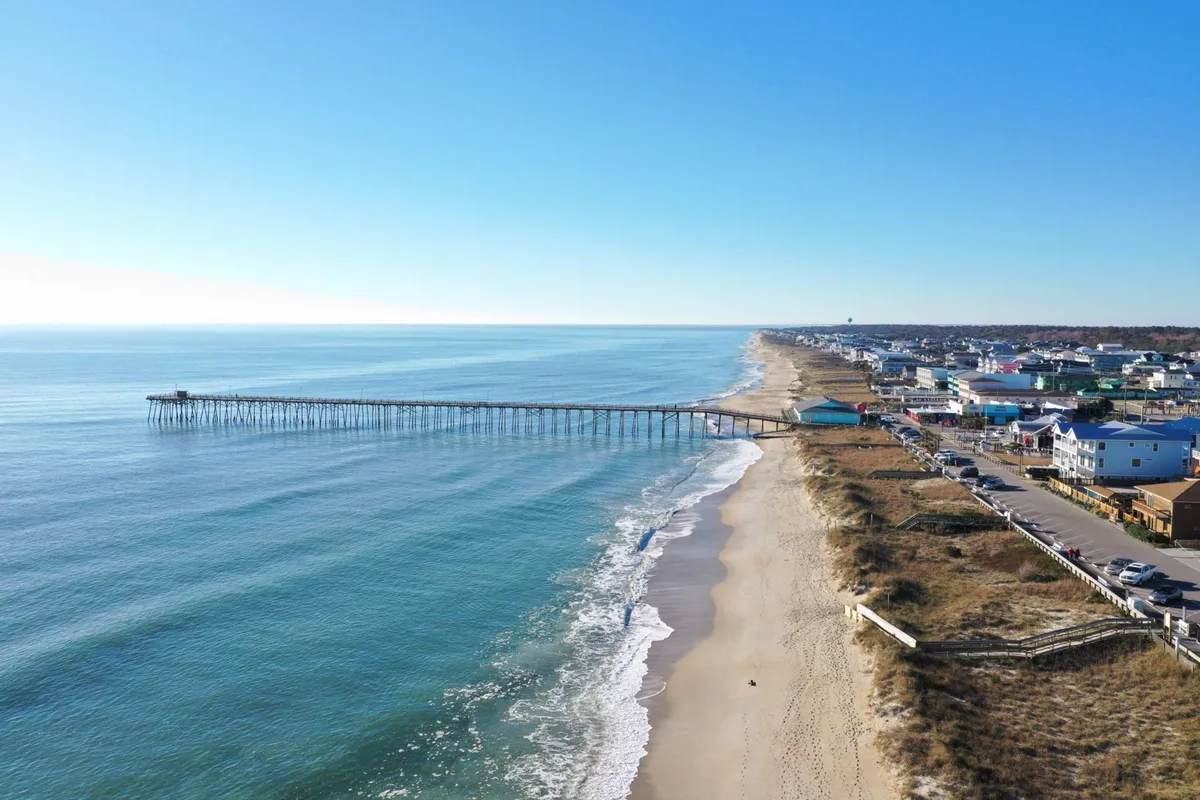 A coastal scene with a pier stretching into the ocean, sandy beach, and charming coastal homes. People can be seen walking along the beach, evoking a sense of community and local life.