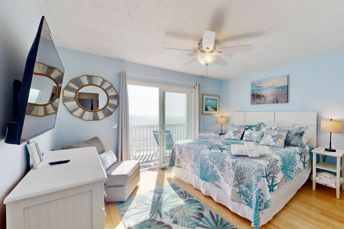 A spacious bedroom featuring a large bed with tropical bedding, sliding glass doors that open to a balcony, a ceiling fan, and decorative mirrors. The atmosphere is serene and tranquil, ideal for rest.