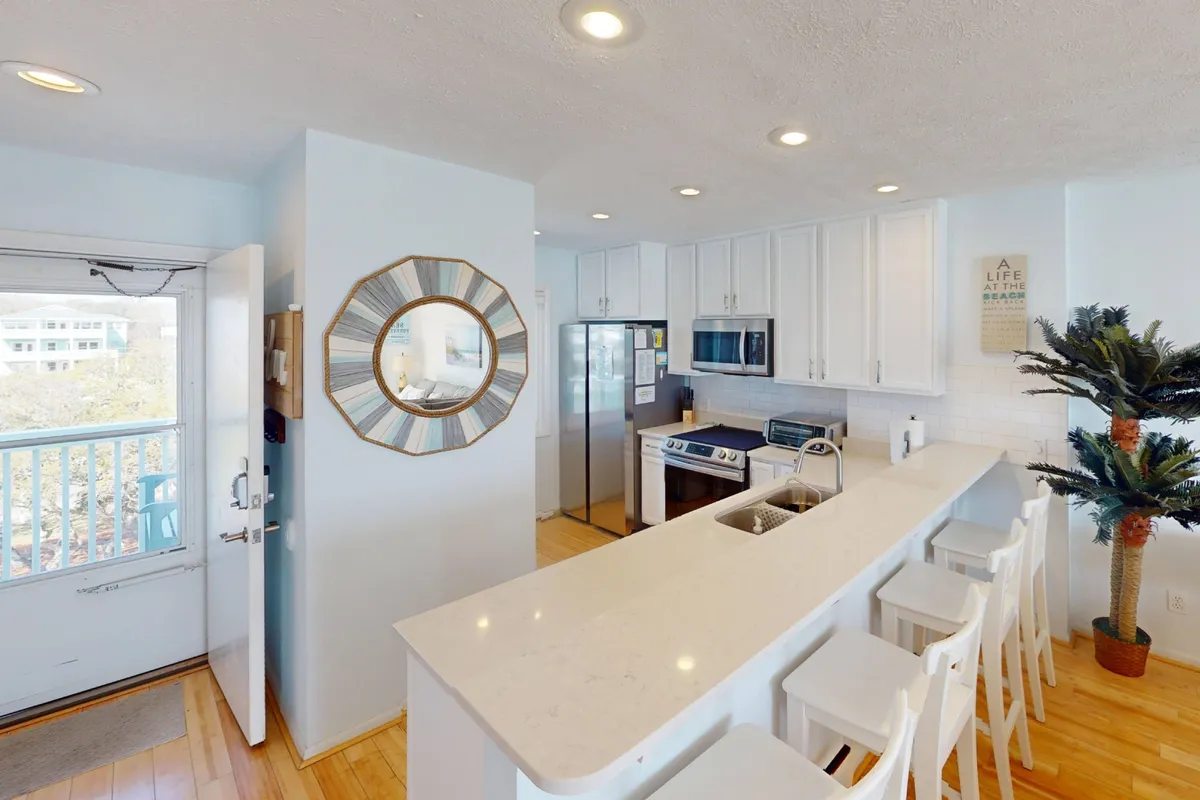A bright kitchen scene featuring a spacious kitchen island, modern stainless steel appliances, a mirror, and a lush palm plant. The design is open and inviting, with ample space for cooking and gathering. The overall mood is cheerful and family-friendly, ideal for preparing meals together or entertaining guests.