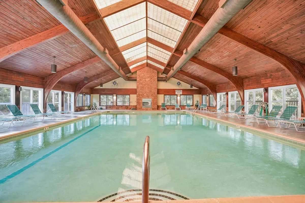 A tranquil indoor swimming pool with wooden beams and large windows letting in natural light. Lounge chairs are arranged along the side of the pool, creating a relaxing environment for year-round swimming and relaxation.