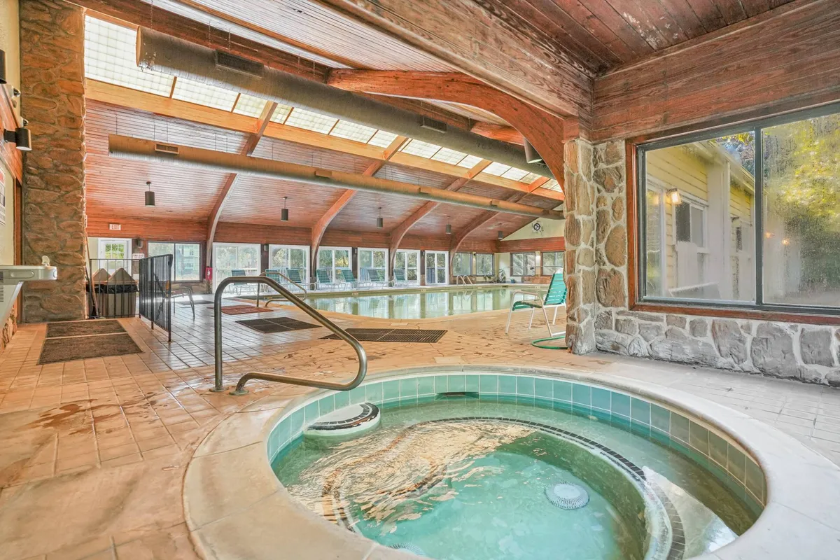 A spacious indoor pool area featuring a large heated pool and a bubbling hot tub, with natural light pouring in through big windows. The inviting setting suggests year-round swimming and relaxation options for families.