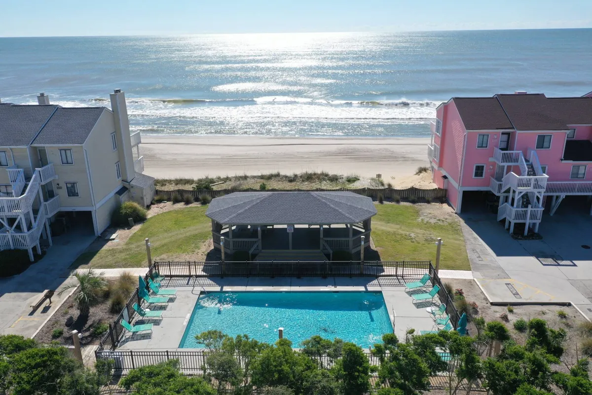 An exterior view of the resort complex, focusing on a sparkling swimming pool with a nearby gazebo, colorful resort buildings, and the ocean in the background. The pool area is tranquil and inviting, set up for both relaxation and play.