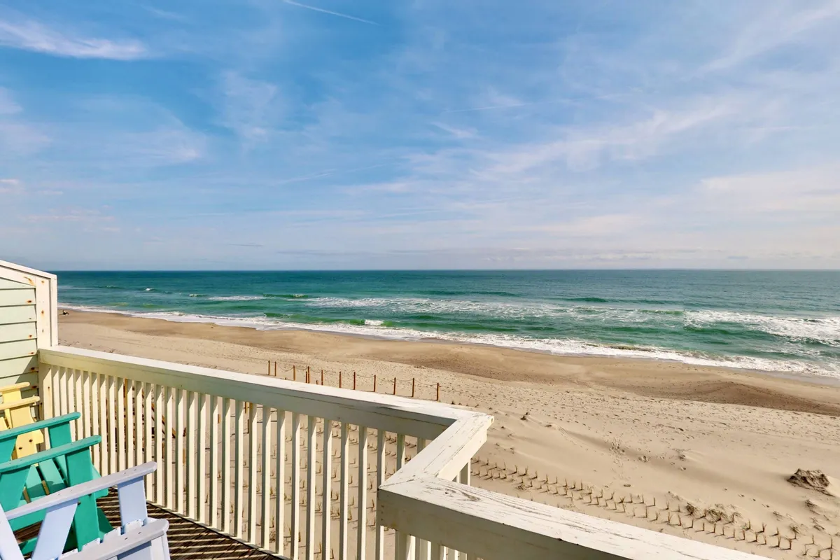 This image captures a serene view from a deck featuring comfortable chairs overlooking the ocean and beach. Soft sunlight illuminates the waves, and the inviting setting is ideal for relaxing, enjoying meals, or intimate conversations while watching the horizon.