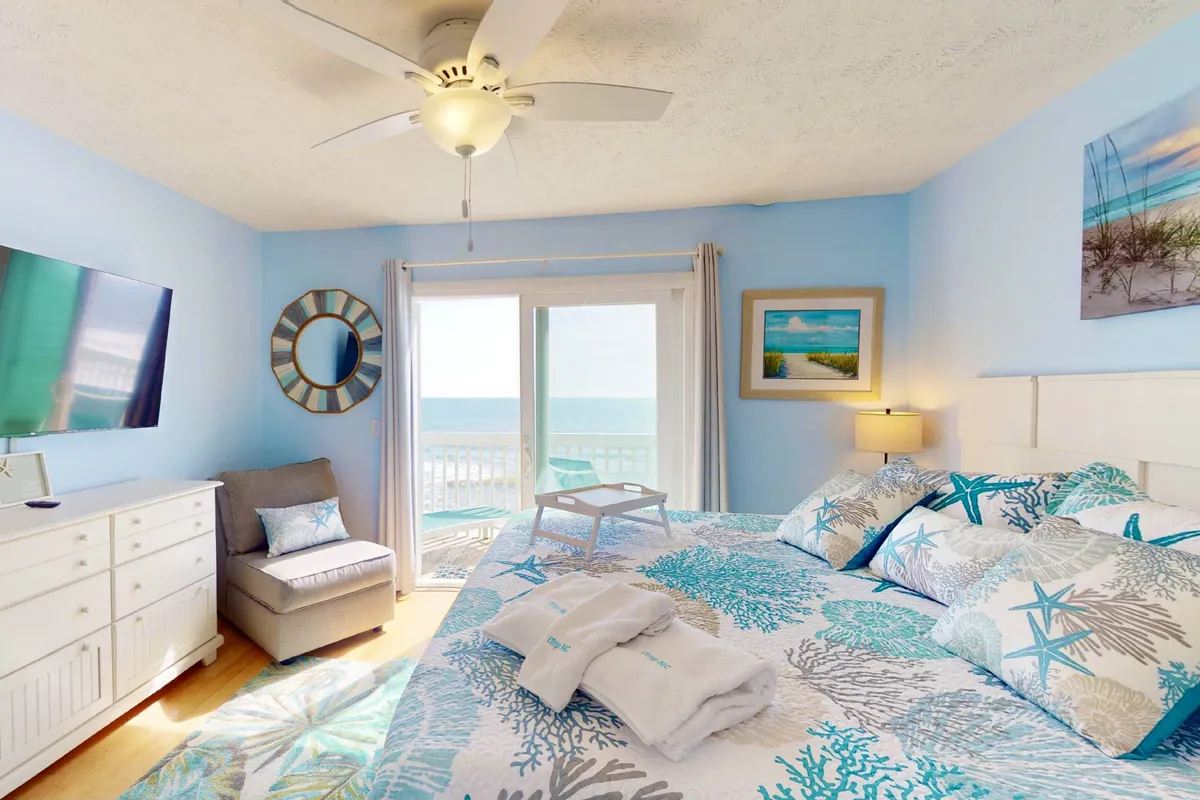 A serene bedroom with colorful bedding, a large window offering an ocean view, a TV on the dresser, and a decorative mirror. Natural light fills the room, making it both inviting and restful.