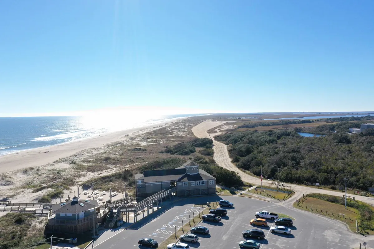 This view showcases the ocean, sandy beach, a boardwalk leading toward the shore, and a parking lot in the background. The scene is lively and inviting, suggesting easy access to beachside activities and coastal exploration.