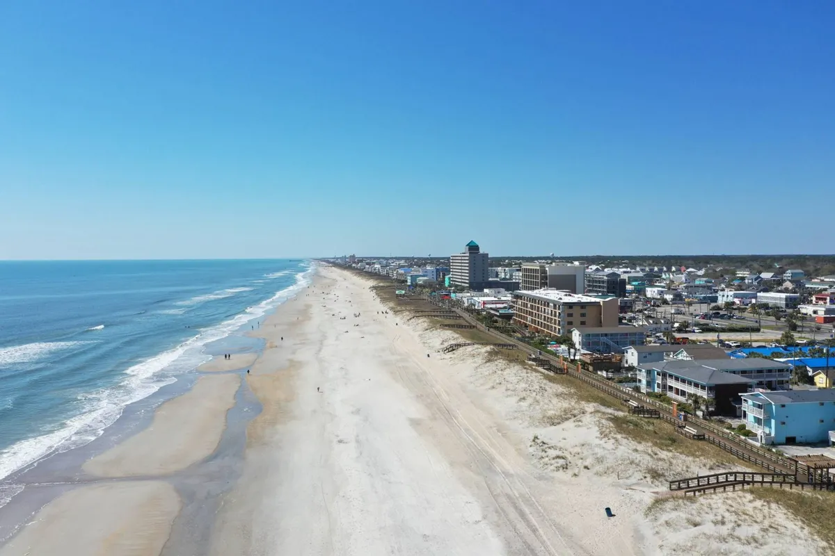 A wide, sandy beach stretches into the distance with gentle ocean waves rolling in. The coastal town skyline is visible beyond the dunes, suggesting close proximity to local amenities. Beachgoers can be seen walking along the shore, swimming, surfing, and sunbathing, all under a relaxed, carefree atmosphere.