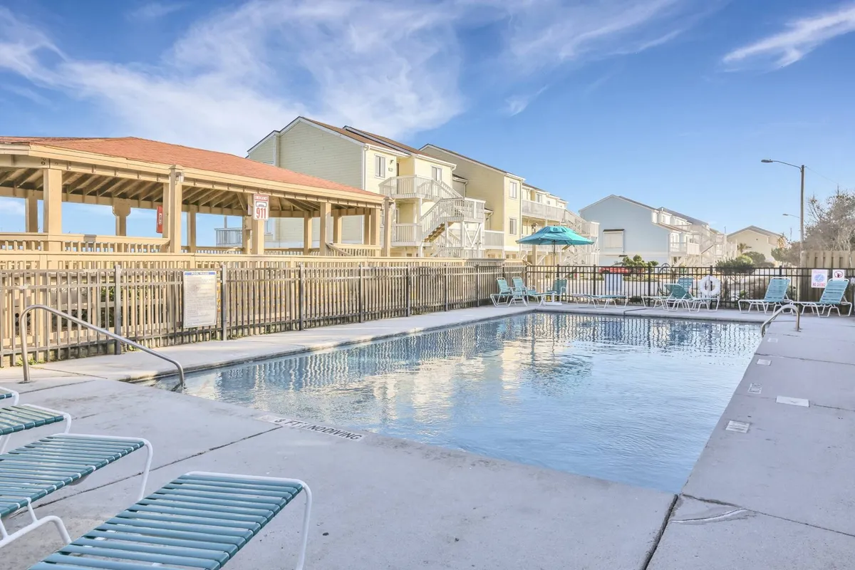 A sparkling outdoor swimming pool flanked by lounge chairs and a covered seating area, with surrounding buildings visible in the background. The pool area is inviting and well-maintained, suggesting a perfect spot for sunbathing, swimming, and socializing during warmer months.