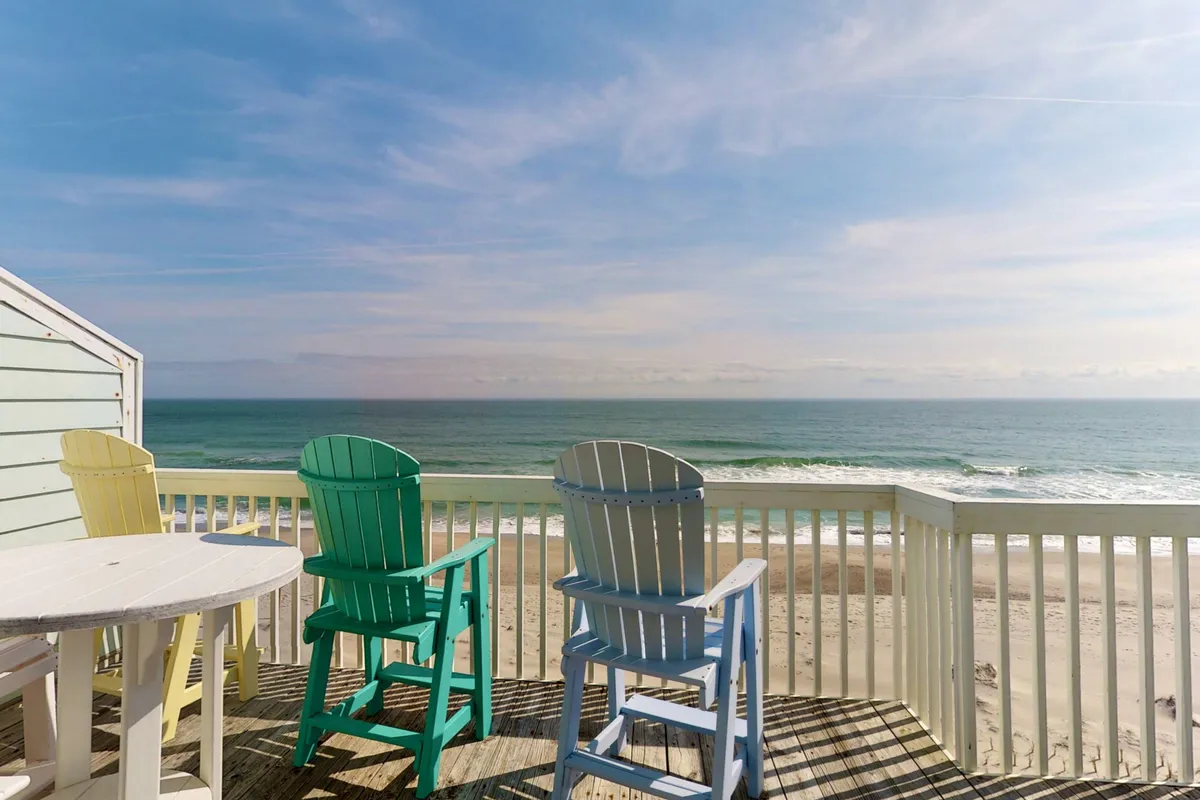 An inviting top-floor deck furnished with colorful Adirondack chairs facing the ocean. The view reveals an endless expanse of blue water and sandy beach beyond the deck railing, suggesting the perfect spot for morning coffee or sunset cocktails while listening to the waves.